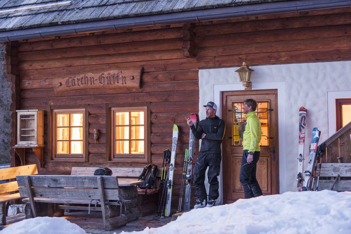 2 Männer stehen vor der Lärchn-Hüttn, an der Wand lehnen einige Paar Ski.