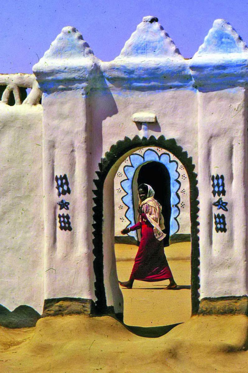 Blick durch ein bunt bemaltes Tor in ein Dorf, eine Frau geht gerade durch die Straßen.