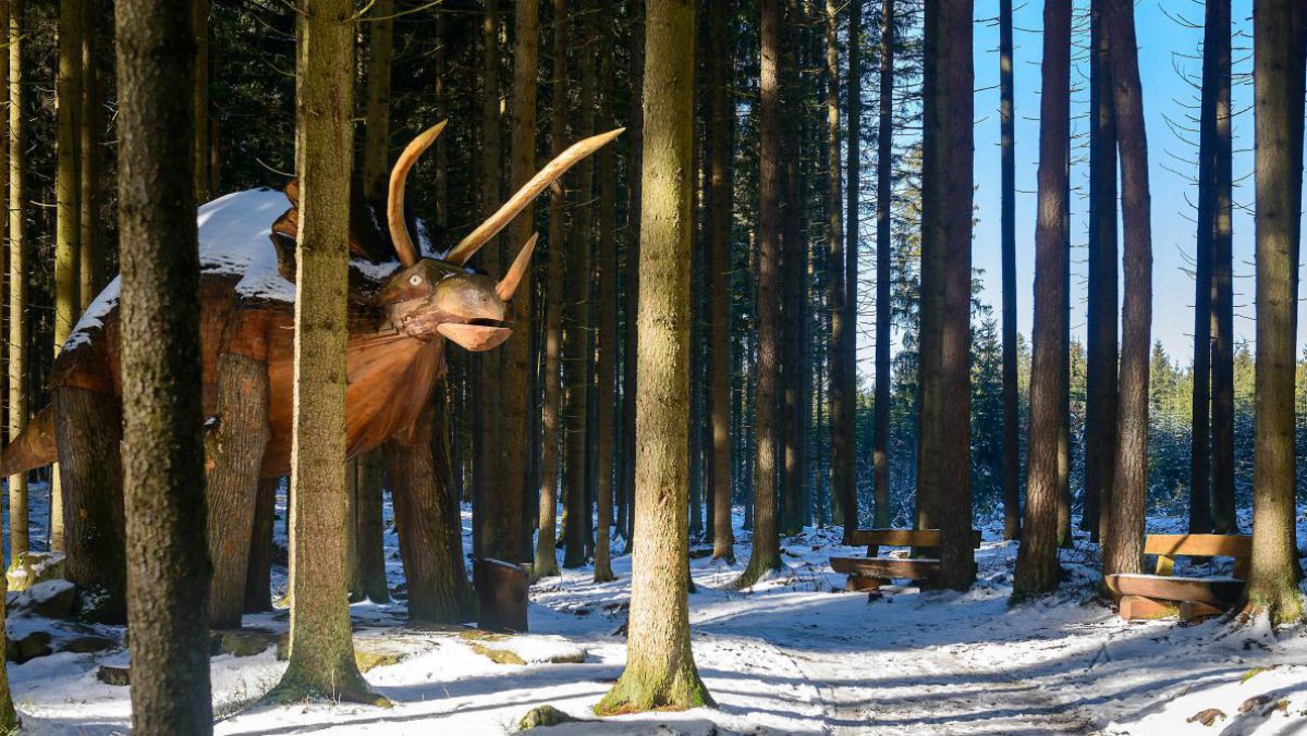 Eine lebensgroße Dinosaurierfigur steht zwischen Baumstämmen am winterlich schneebedeckten Erlebnisweg Moorwald.