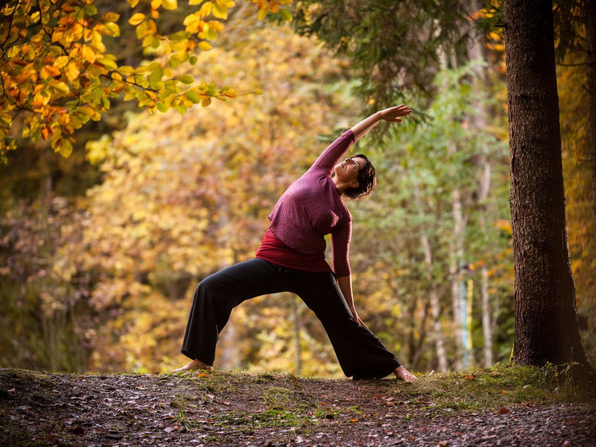 Eine Frau steht in einer Yoga-Position auf einem Weg in einem herbstlich gefärbten Mischwald.
