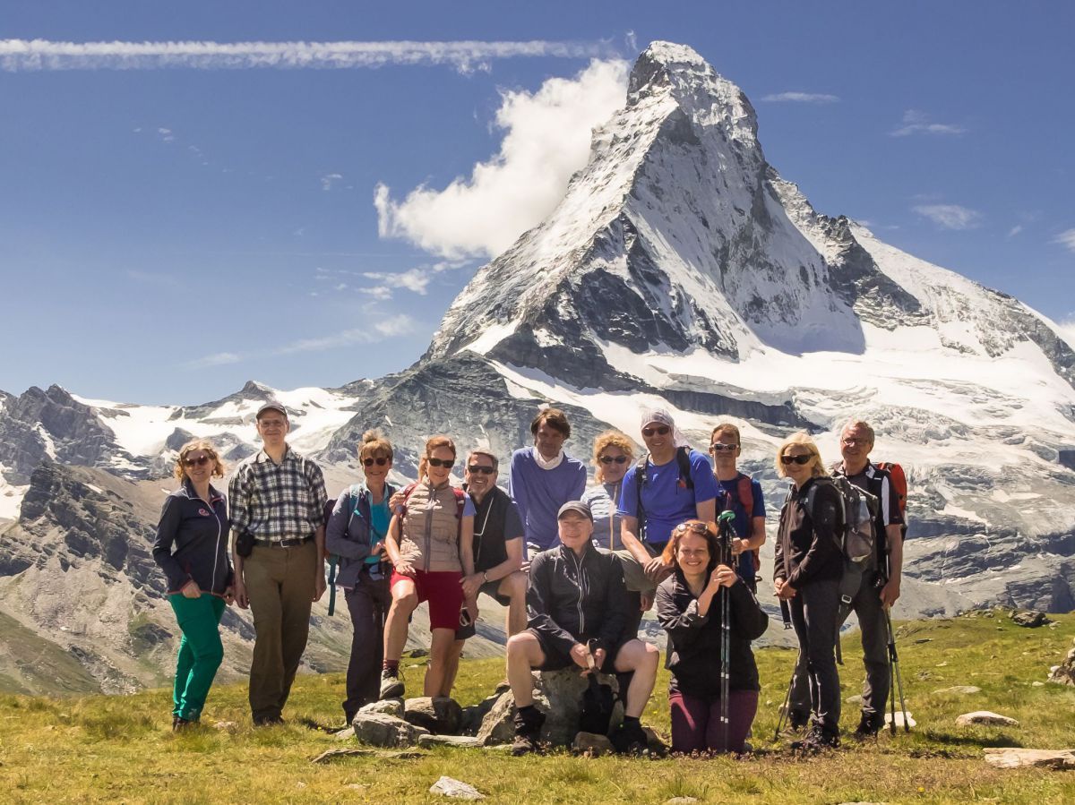 Eine größere Gruppe Wanderer auf dem mit Gras bewachsenen Hochplateau eines Berges, im Hintergrund ein noch höherer, schneebedeckter Gipfel vor einem blauen, leicht bewölkten Himmel.