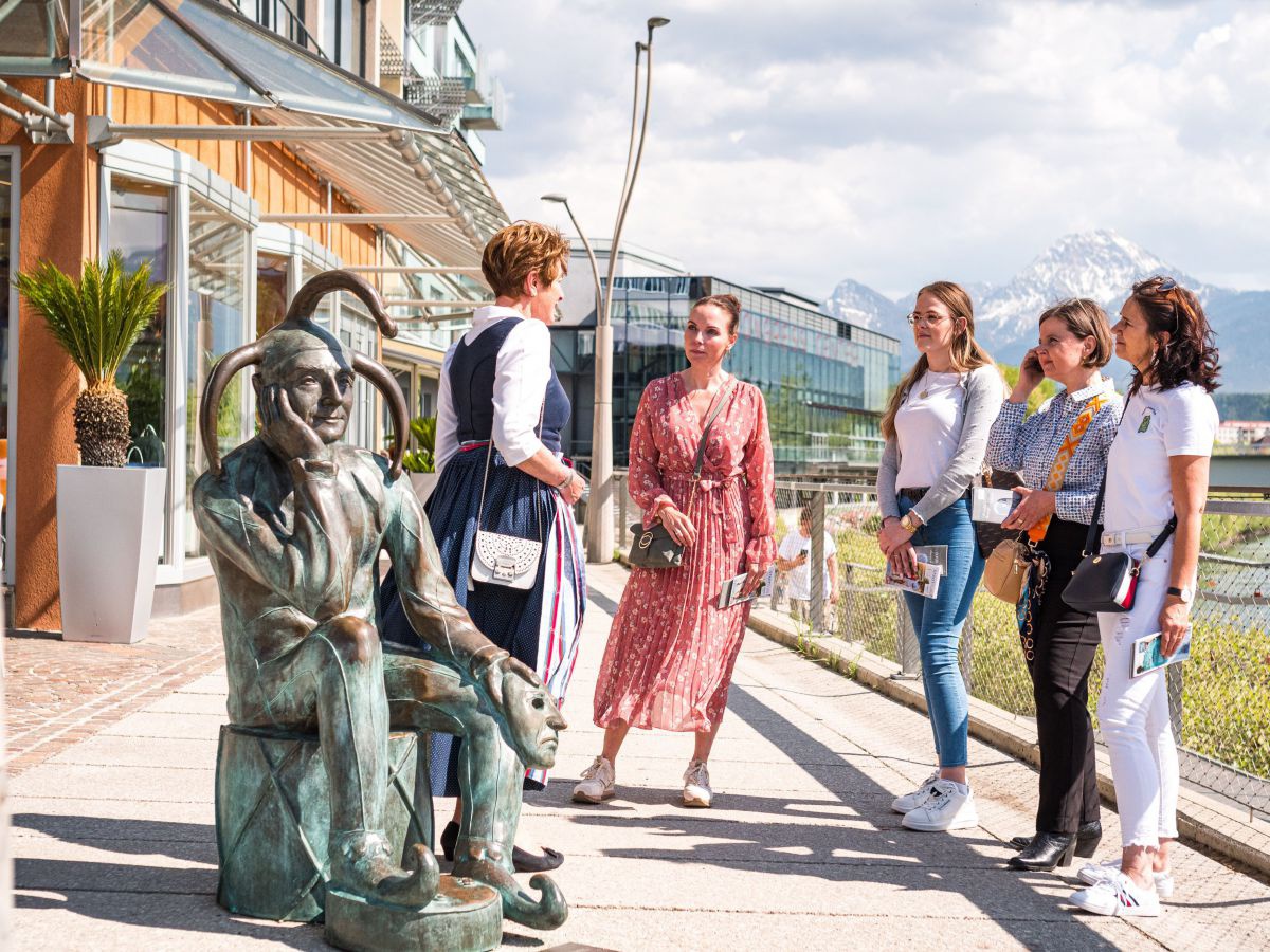 Vier junge Frauen  und eine Führerin im Dirndl bei einer Stadtführung in Villach auf der Uferpromenade am See. Im Vordergrund die Bronzestatue eines sitzenden Till Eulenspiegels.