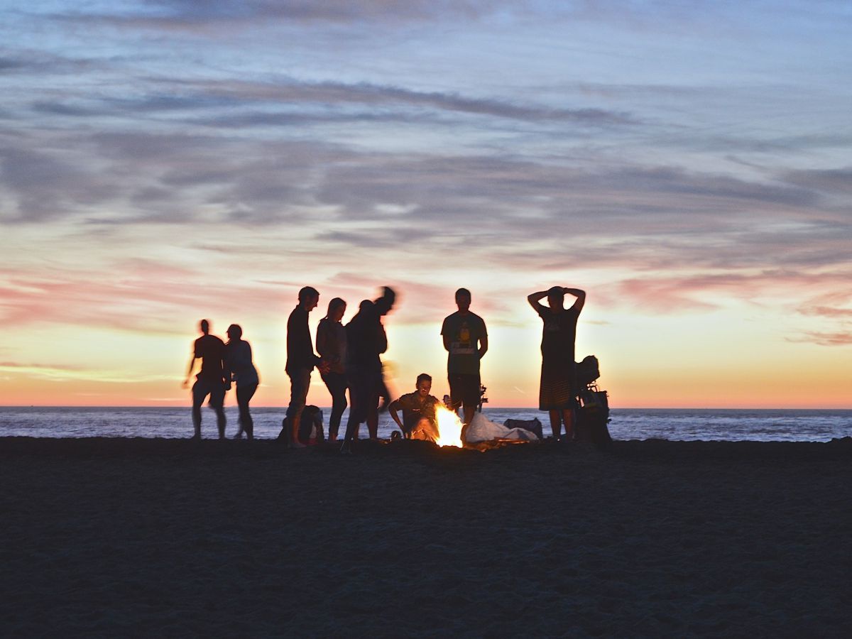 Eine Gruppe Menschen bei Lagerfeuer am Strand nach Sonnenuntergang. Der Himmel über dem Meer bietet ein eindrucksvolles Farbenspiel in Gelb-, Rosa- und Blautönen.