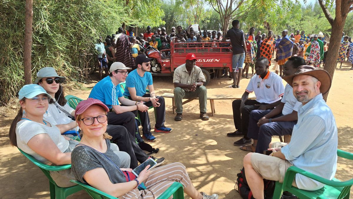 Reisegruppe sitzt mit afrikanischen Männern im Kreis, dahinter eine Gruppe von Menschen hinter einem kleinen Truck.