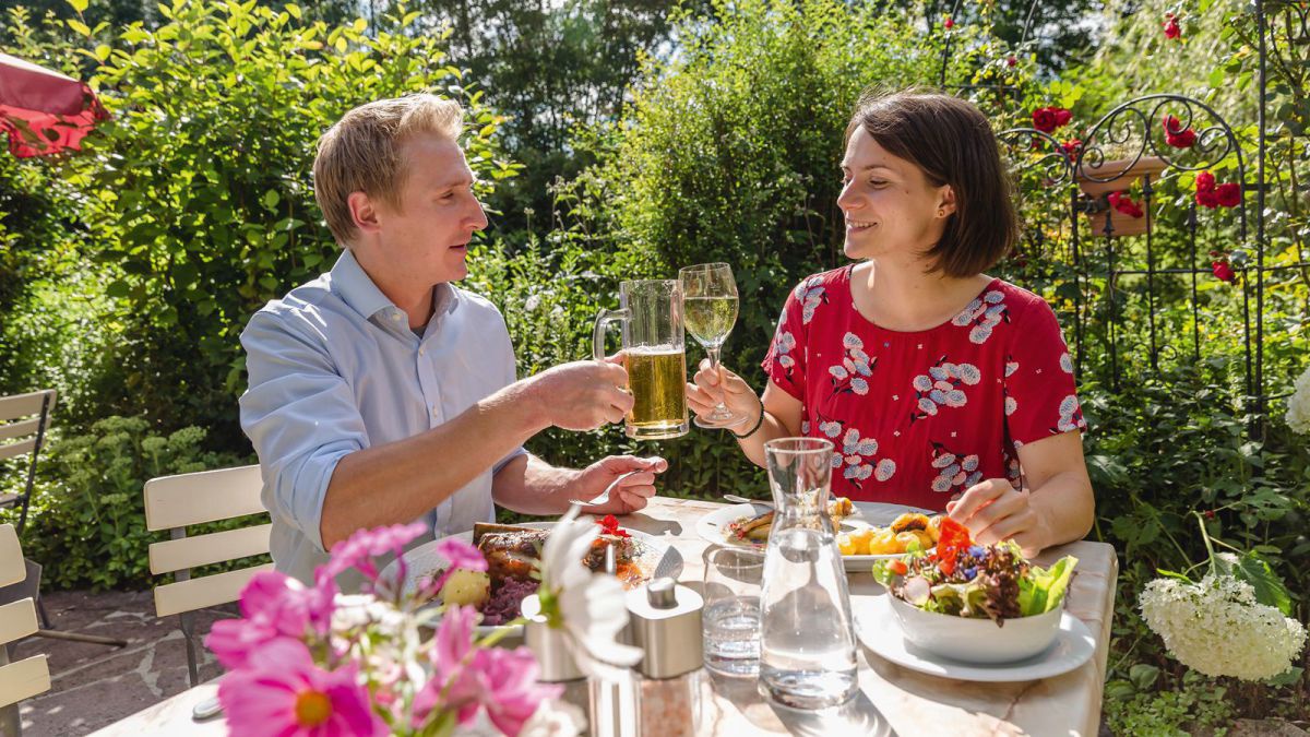Mann mit einem Krügerl Bier und Frau mit einem Glas Wein prosten sich zu. Auf dem Tisch sind mehrere Speisen zu sehen und eine Karaffe Wasser.