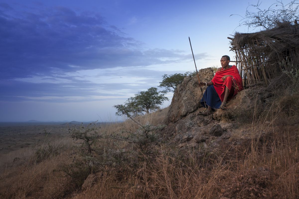 Maasai sitzt bei einem Felsen mit einem Stab in der Hand.