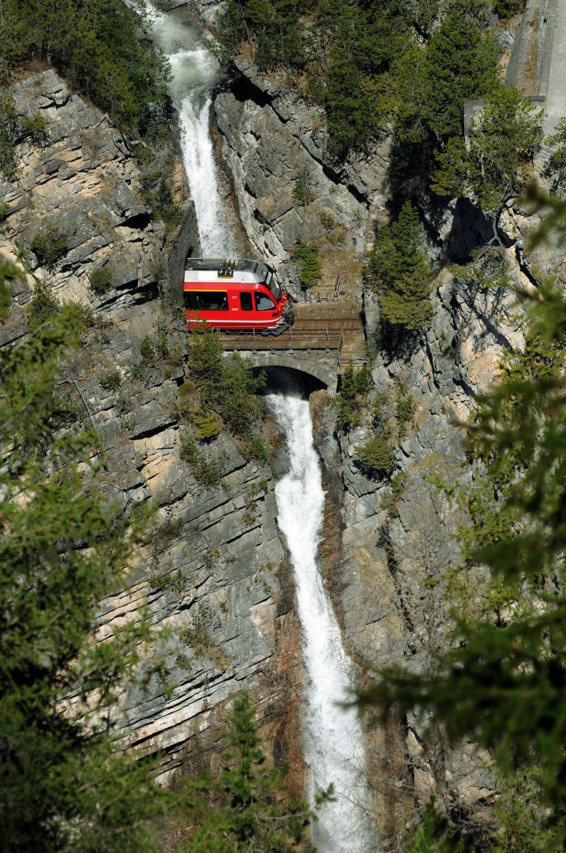 Zug fährt auf ganz kurzer Brücke zwischen 2 Tunnels über einen Wasserfall.