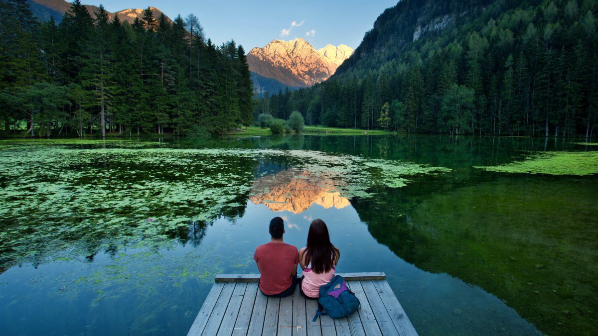 Paar sitzt uf einem Steg und schaut über den See zu einem mächtigen Berg, der von der Sonne beleuchtet ist.