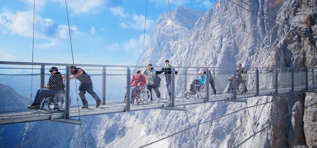 Gruppe von Rollstuhlfahrern mit Begleitung auf einer Hochseilbrücke zwischen den Felsen.