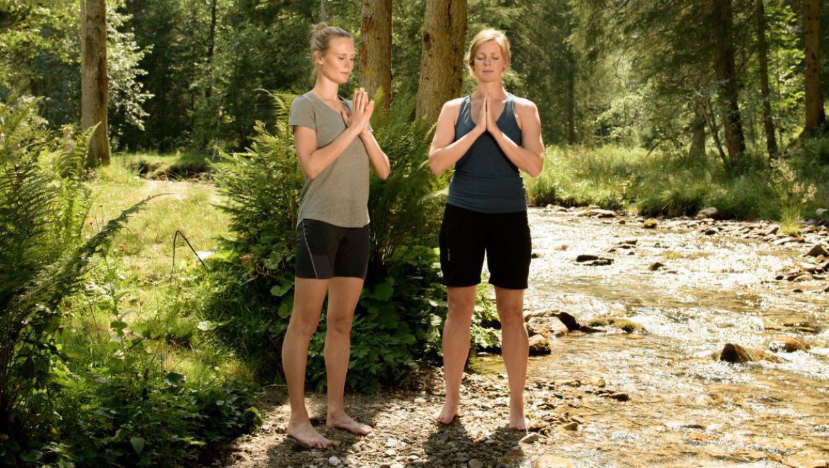 2 Frauen meditieren stehens im Wald am Ufer eines Baches.