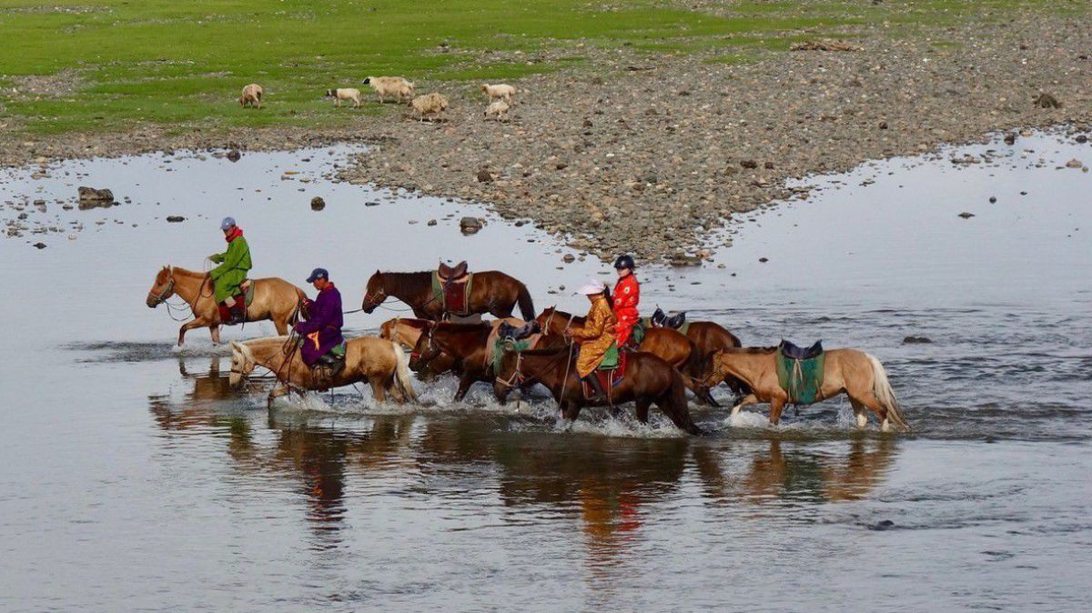 Gruppe von 4 Personen mit 7 Pferden in der mongolischen Steppe reitet durch eine Wasserstelle.