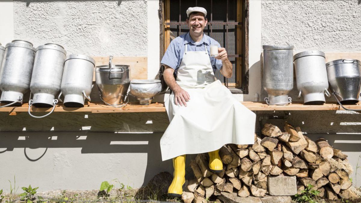 Mann mit einem Glas Milch sitzt auf einer Brett zwischen Milchkannen.