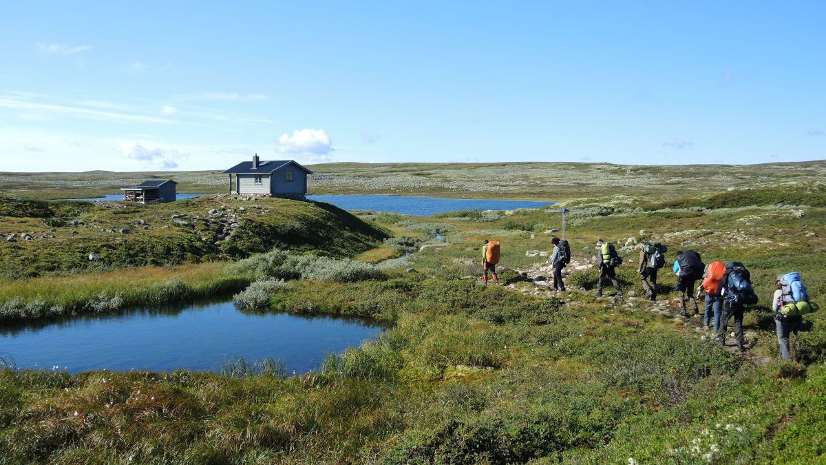 Eine Gruppe wandert auf einer Hochebene zwischen 2 kleinen Seen in Richtung einer kleinen Hütte.