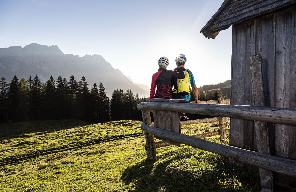 Paar sitzt auf einem Holzzaun vor einer Hütte und schaut in die Morgensonne.