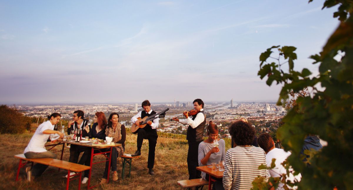 Heurigentisch mit Gästen, 2 Musiker stehen mein Tisch und spielen, dahinter das Panorama der Stadt Wien.