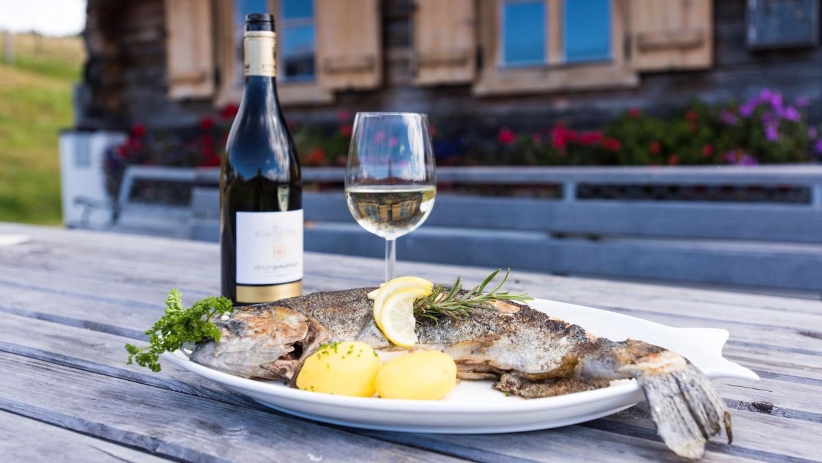 Auf einem Holztisch im Freien vor einem Holzhaus stehen einTeller mit Fisch, eine Flasche und ein Glas Wein 