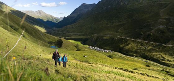 3 Personen wandern über grüne Wiesen durch ein Tal, im Hintergrund Berge.