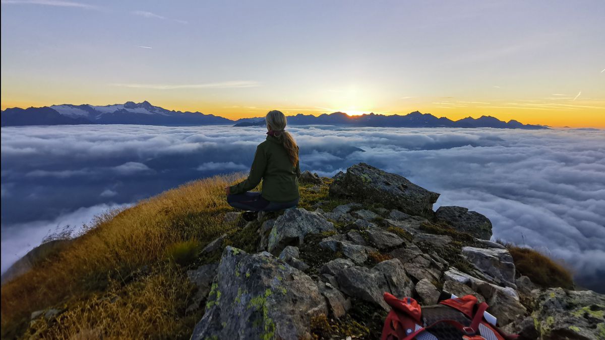 Irmgard Wibmer sitzt auf einen Berggipfel bei Sonnenaufgang. Unter ihr ein Nebelmeer.