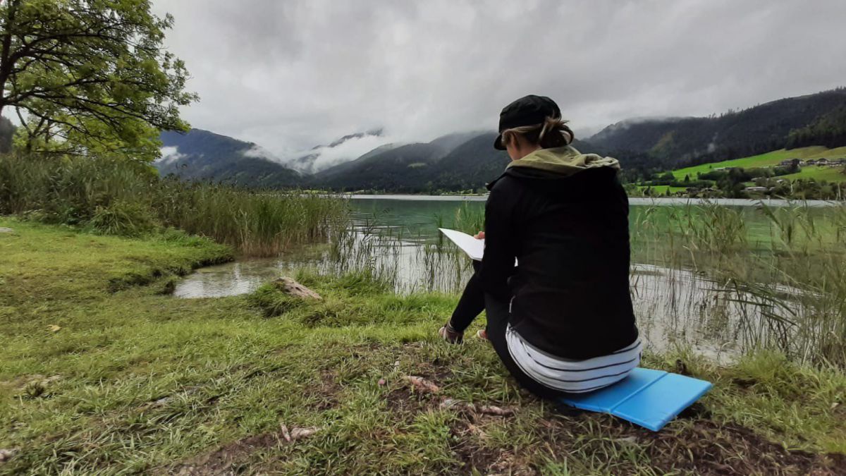 Eva Maria Resch sitzt im Gras am Ufer des Weissensees und zeichnet.