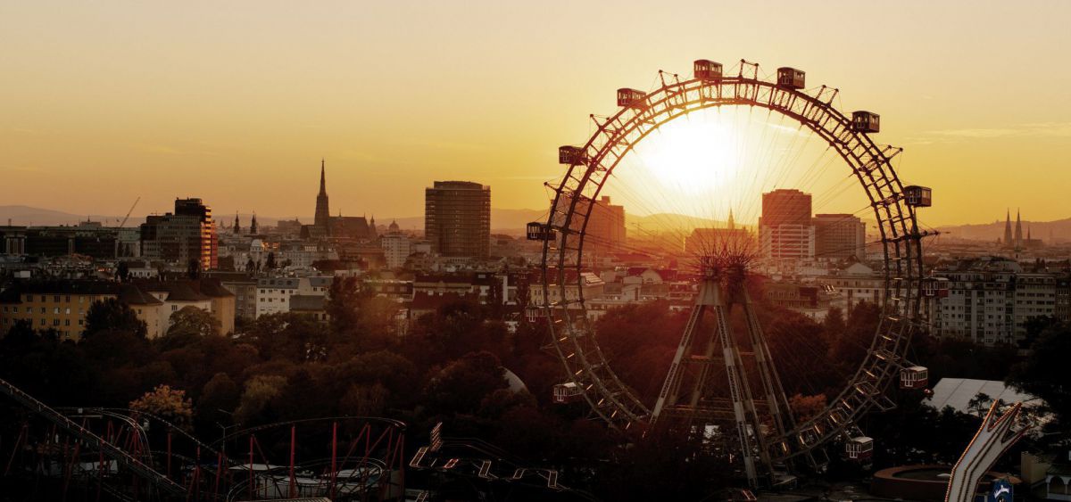 Blick auf die untergehende Sonne, im Vordergrund das Riesenrad, dahinter die Stadt.