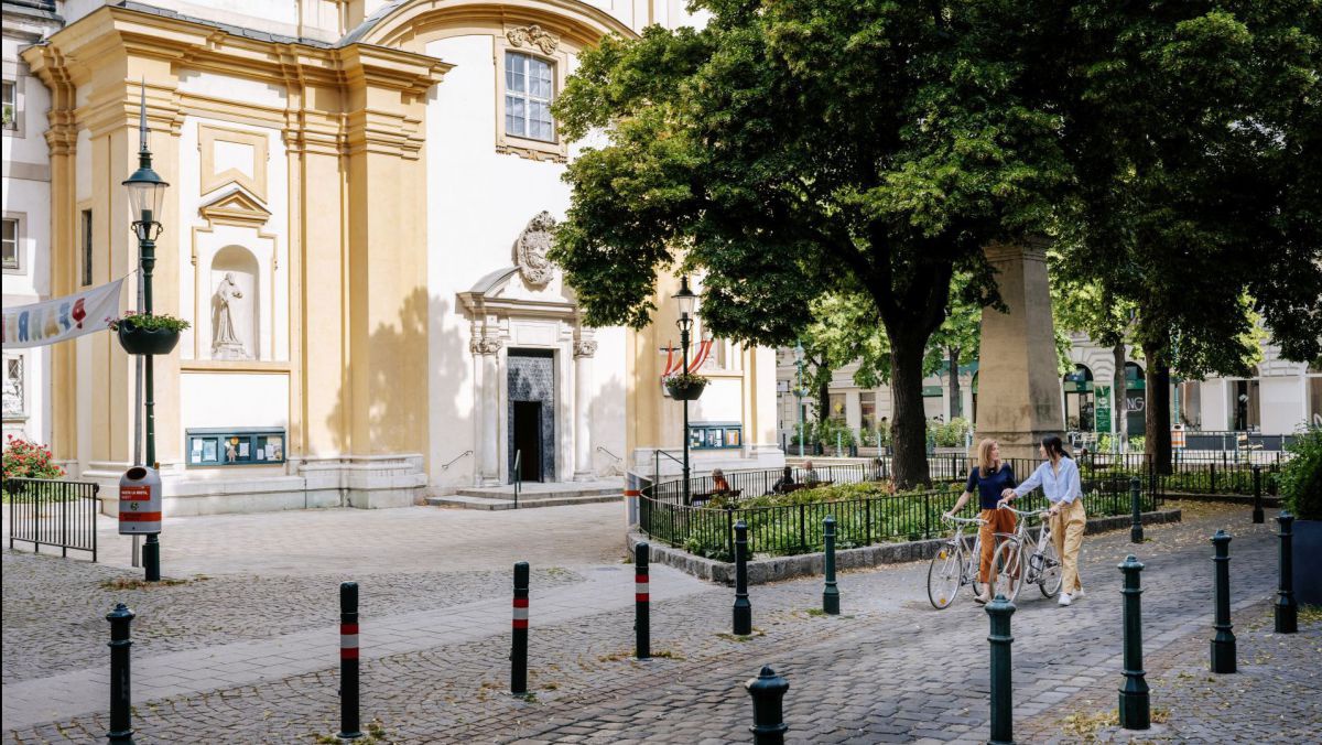 2 Frauen schieben ihre Fahrräder auf einer mit Kopfsteinpflaster ausgelegten Gasse vor einer Kirche.