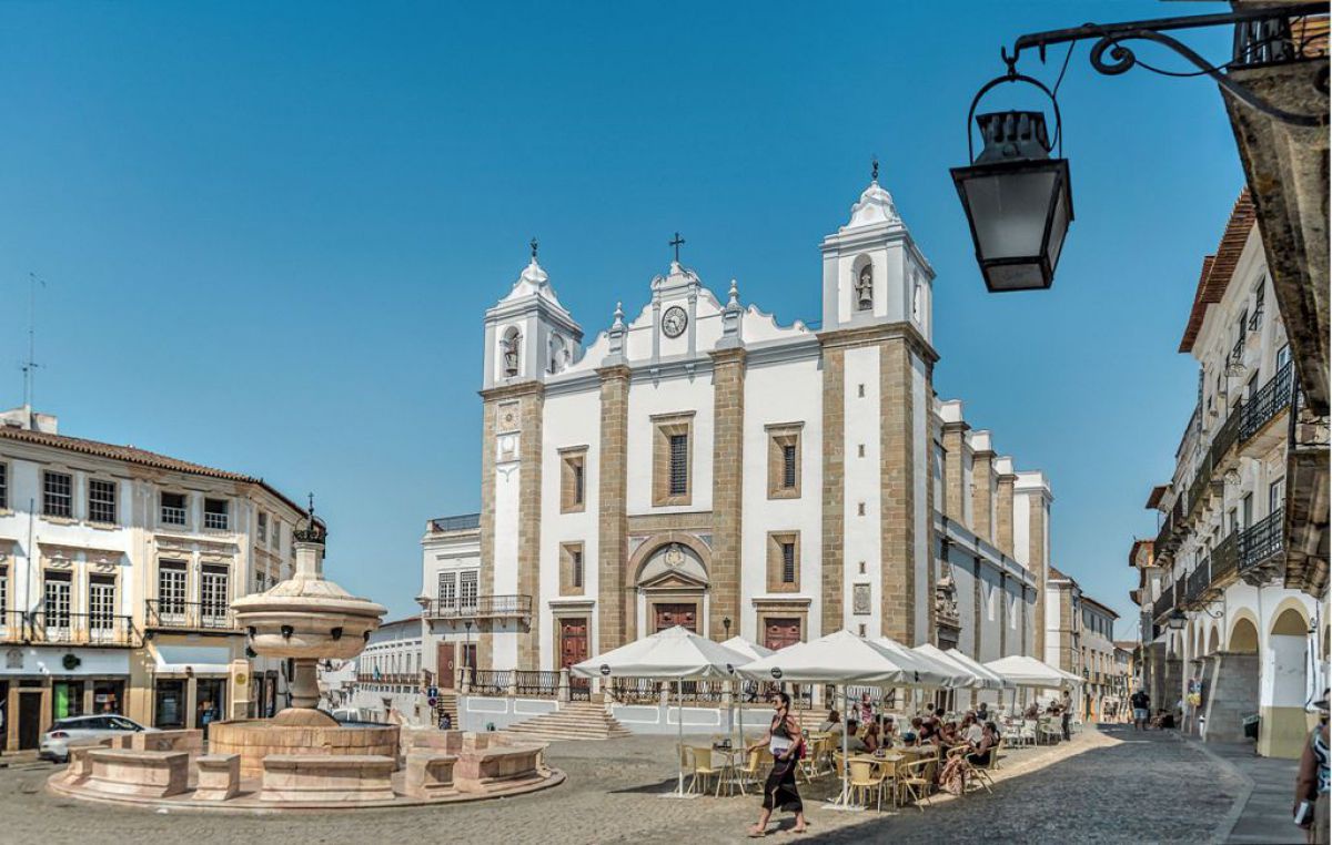 Kathedrale aus weißem und rötlichem Marmor, davor Hauptplatz mit Brunnen und Tischen und Sesseln unter Sonnenschirmen.