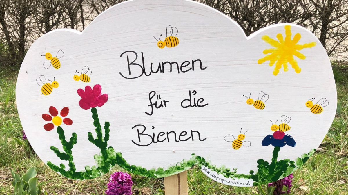 Eine von Kindern bemalte Holztafel in Form einer Wilke. Darauf steht: Blumen füt die Bienen. Und es sind Blumen, Bienen und die Sonne draufgemalt.