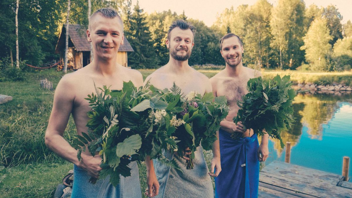 3 Männer mit nacktem Oberkörper und Büchen aus Zweigen am Steg eines kleinen Sees.
