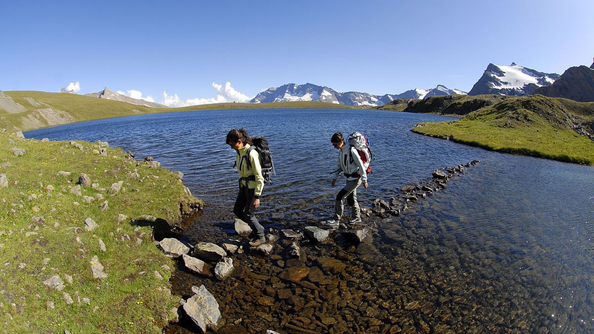 2 Wanderer überweren über Steine einen seichten See im Hochgebirge.