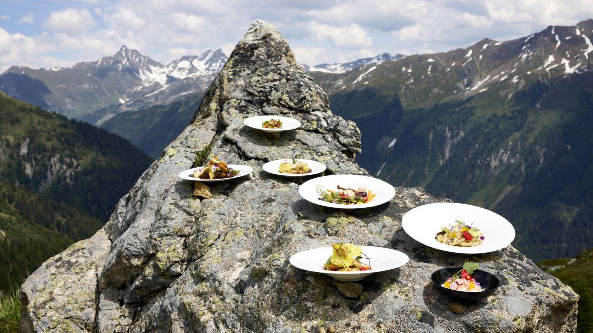 Sieben Teller mit schön angerichteten Speisen stehen auf einem Fels vor beeindruckendem Bergpanorama.