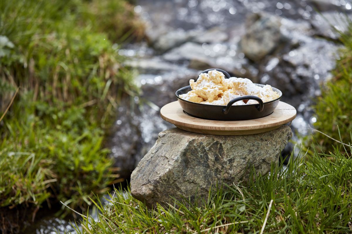 Eine gusseiserne Pfanne mit Schmarren auf einem Holzbrett steht auf einem Stein vor einem Bächlein.