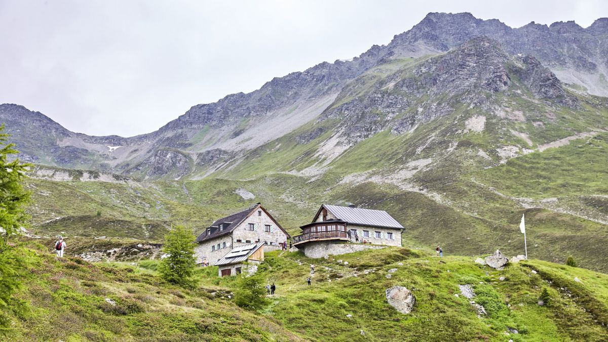 Ein Blick von etwas unterhalb auf die beiden Gebäude der Friedrichshafener Hütte. Dahinter erhebt sich schroff die Bergspitze.