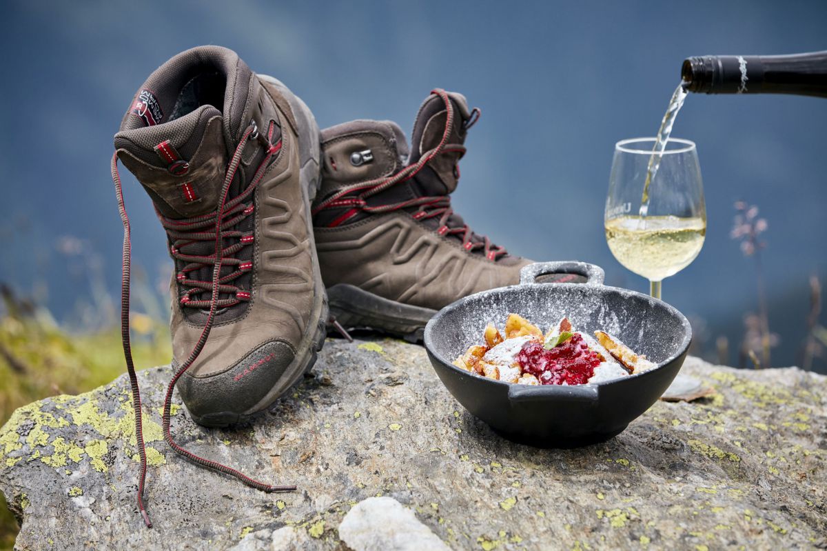 Auf einem Stein stehen ein Paar Wanderschuhe, eine Mehlspeise in einer Schüssel und ein Glas Wein, in das gerade eingeschenkt wird.