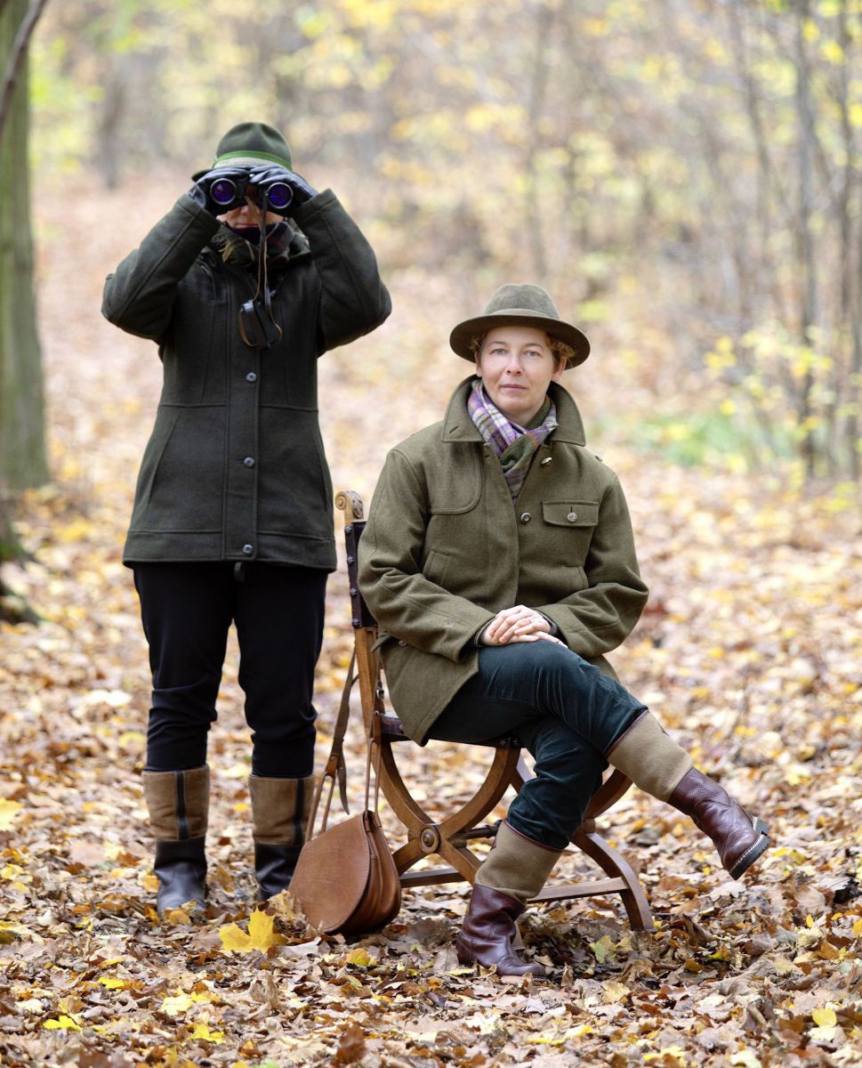 Zwei Jägerinnen im Lodenmantel, Stiefeln und Hut. Eine sitzt auf einem Sessel, die andere blickt mit einem Fernrohr in die Kamera. Rundherum Herbstwald.