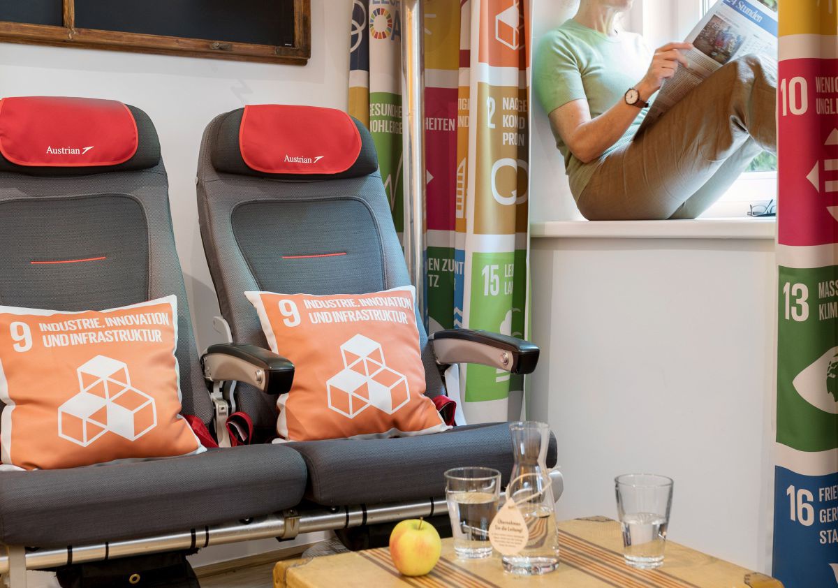 Zimmer mit alten Sitzen aus einem AUA Flugzeug, dasin Pölster mit SDG 9. Am Vorhang sind die Icons aller SDG's abgebildet.