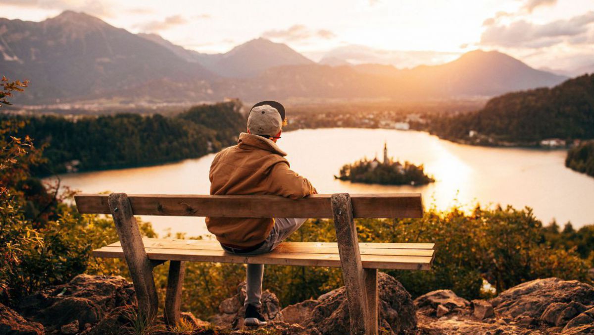 Ein Junger Mann mit Schirmkappe sitzt entspannt auf einem hochgelegenen Bank mit Blick auf den See bei Sonnenuntergang