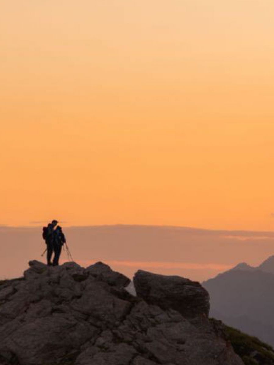 Zwei Wanderer stehen am Gipfel eines Berges im zart-rosa-orangen Licht des Sonnenaufgangs