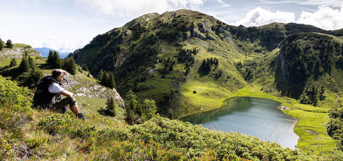 Ein Mann in Wanderhose sitzt im Heidekraut und blickt hinunter auf einen See inmitten zart bewaldeter Berge, es ist sonnig und leicht bewölkt.