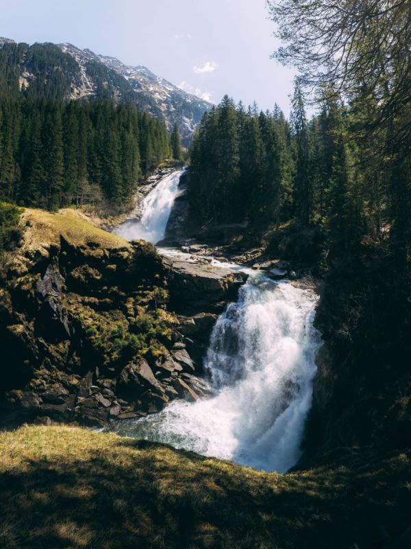 Zwischen hohen Fichten tost ein Wasserfall über zerklüftetes Gestein, im Hintergrund ein Berggipfel vor blauem Himmel an einem sonnigen Tag.