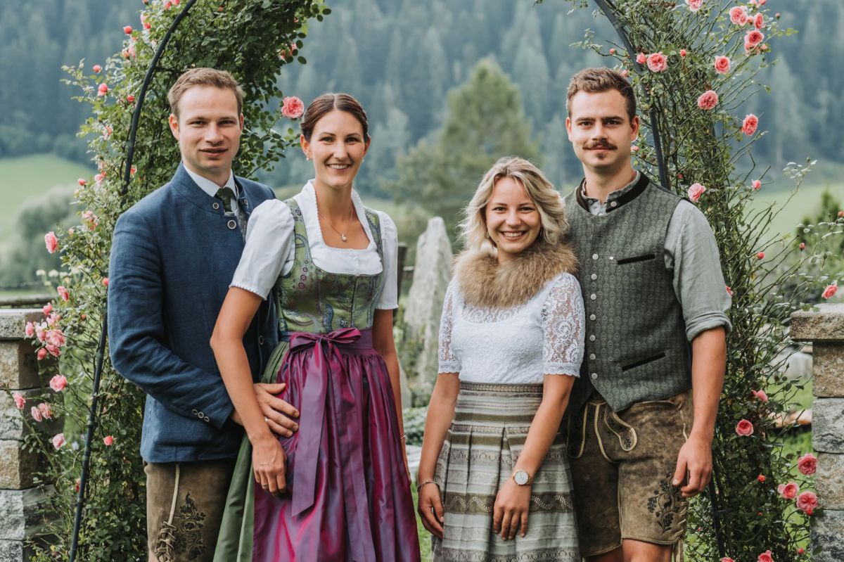 Zwei Männer und zwei Frauen in Tracht gekleidet, vor einem Rosenbogen posierend.