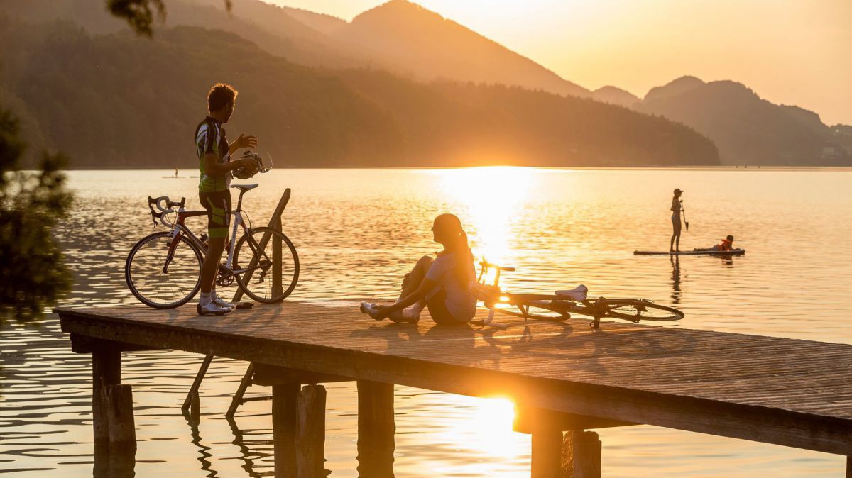 Zwei Radfahrer machen Pause auf einem Steg am Seeufer, die untergehende Sonne spiegelt sich im Wasser, im Hintergrund Berge.
