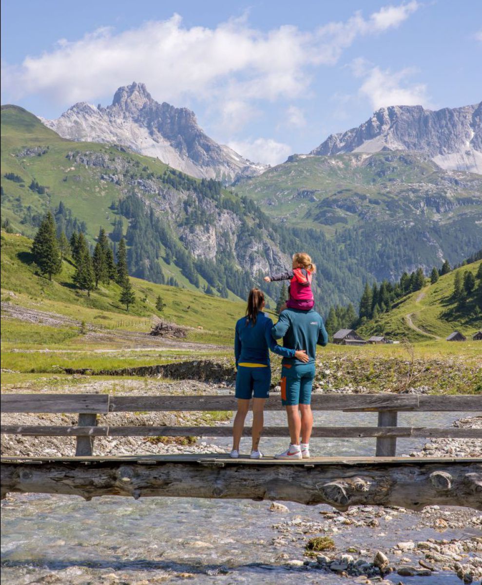 Ein Paar steht auf einer hölzernen Brücke über einem Gebirgsbach, der Vater trägt ein kleines Mädchen auf den Schultern, alle drei blicken in die gewaltige Kulisse aus Berggipfeln und bewaldeten Hängen, der Himmel ist blau mit ein paar weißen, lockeren Wo