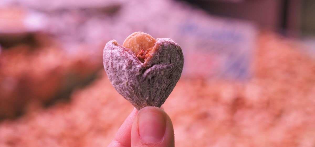 Ein kleines Herz, aus einer getrockneten Feige und einer Mandel zusammengesetzt, wird von einer weiblichen Hand zwischen Daumen und Zeigefinger ins Bild gehalten. Im Hintergrund sieht man verschwommen einen Markttisch mit unzähligen anderen Früchten. Die 
