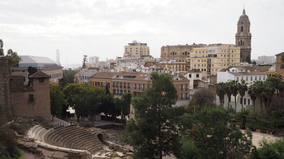 Blick auf das historische Zentrum von Málaga: Reste eines römischen Amphiteaters umringt von hohen Bäumen, im Hintergrund ragt der Turm der Kathedrale zwischen den Stadthäusern empor.