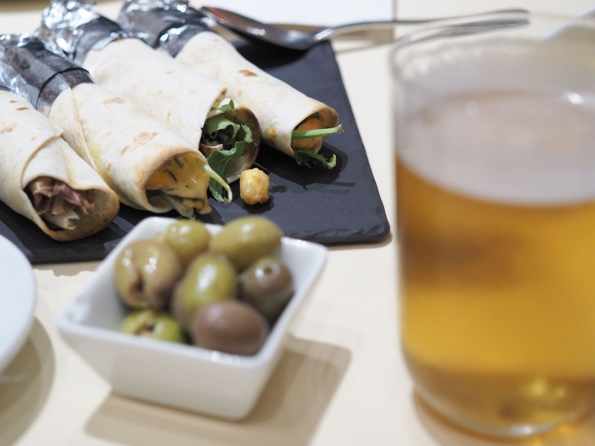 Ein Glas Bier im Vordergrund, links daneben ein Schälchen mit Oliven, dahinter auf einer Schieferplatte angerichtet vier gefüllte, dünne Fladenbrote.