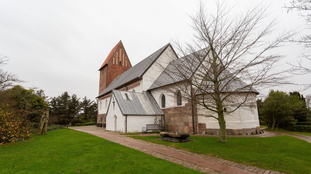 Eine gut erhaltene kleine Kirche aus dem 13. Jahrhundert mit schlichtem, rotgemauertem Türmchen, eingesäumt von einem Weg aus roten Ziegelsteinen, die nass glänzen vom Regen, dazwischen sattgrüne Grasfläche. Der Himmel ist trüb, die Bäume tragen kein Laub