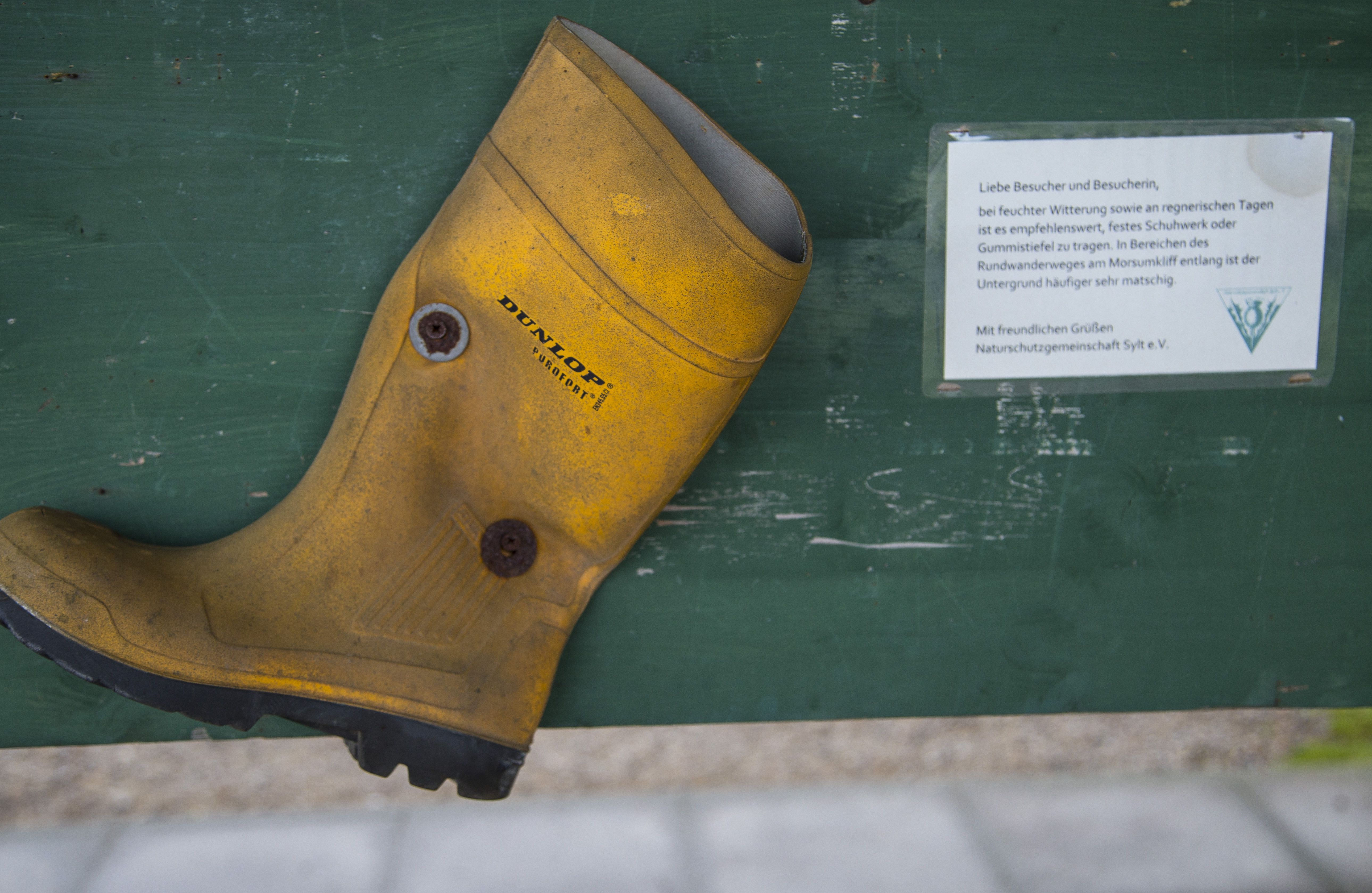 Ein gelber Gummistiefel der Marke Dunlop, an eine grüne Tafel genagelt. Daneben ein Hinweis der Naturschutzgemeinschaft Sylt, den Rundwanderweg am Morsunkliff bei feuchter Witterung vorzugsweise mit Gummistiefeln zu begehen.