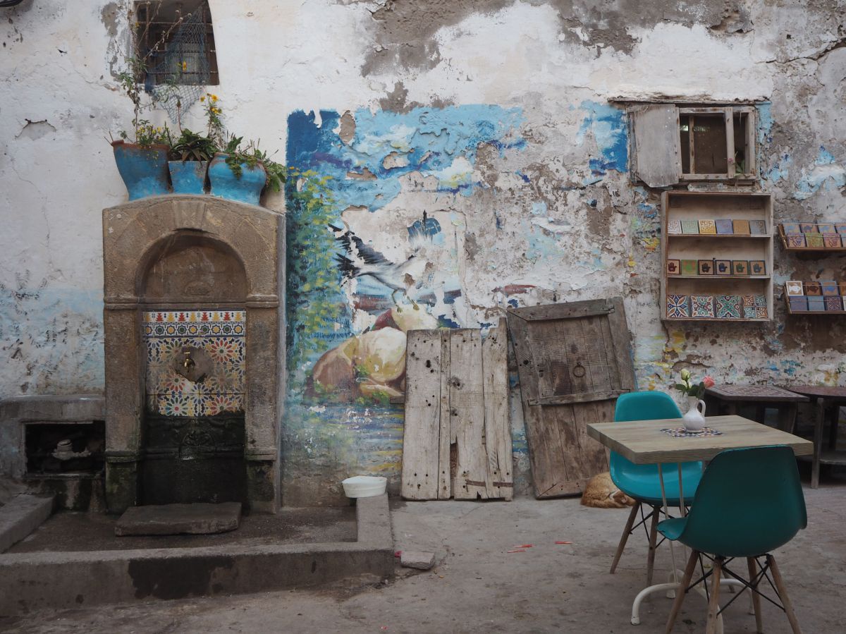 In einem Hinterhof mit mosaikverzierter Bassena steht ein Bistrotisch mit zwei türkisen Stühlen. Die Farbe der Wandbemalung blättert ab, bunte Mosaikfliesen auf alten Holzregalen, türkise Blumentöpfe.