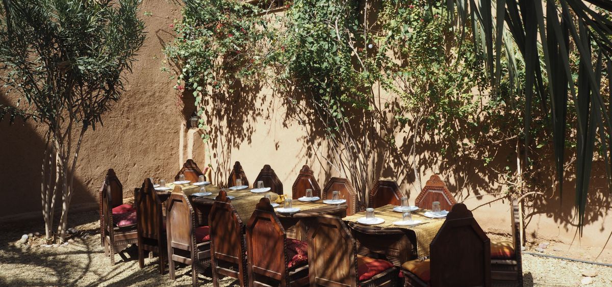 Eine gedeckte Tafel in einem sonnigen Innenhof zwischen sandfarbenen Hausmauern. Tisch und Stühle aus kunstvoll gearbeitetem Holz im marokkanischen Stil, die Fächer der schattenspendenden Palmen hängen ins Bild.