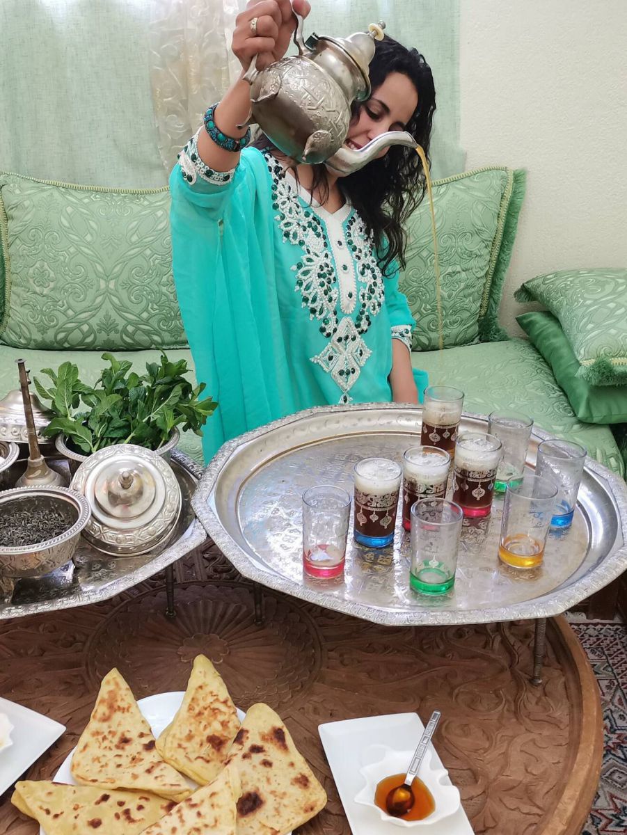 Eine Frau im türkisen, reich bestickten Kleid sitzt auf einem gemusterten, mintgrünen Sofa und schenkt mit gestrecktem Arm aus der sehr hochgehaltenen, silbernen Kanne Tee in bunt verzierte Gläser auf einem Silbertablett...